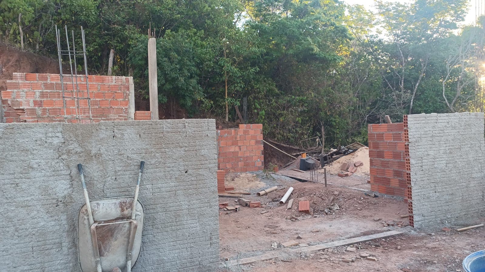Paredes de tijolos em levantamento, com carrinho de mão e materiais no canteiro.
